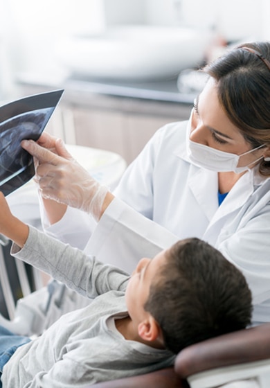 Une dentiste en blouse blanche et masque de protection montre une radiographie dentaire à un jeune garçon assis dans un fauteuil dentaire.